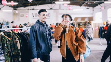 Flohmarkt mit Jugendlichen – Zwei junge Menschen lächeln in einer Halle, umgeben von Kleiderständern und Besuchern.