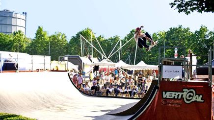 Skateboarder bei Sprung im Freien – Skater führt einen Trick auf einer Rampe vor, im Hintergrund Zuschauer und Stände.