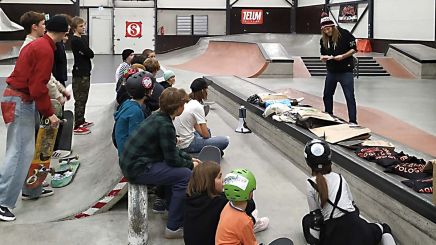 Skateboard-Workshop in der Halle – Gruppe Jugendlicher mit Helmen und Skateboards hört einem Trainer im Skatepark zu.